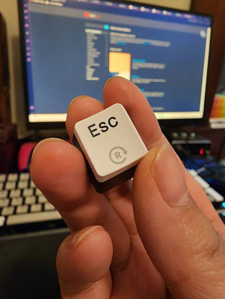 A hand holding the escape keycap from a Hexgears GK735 keyboard, showing the legend on the top of the keycap.