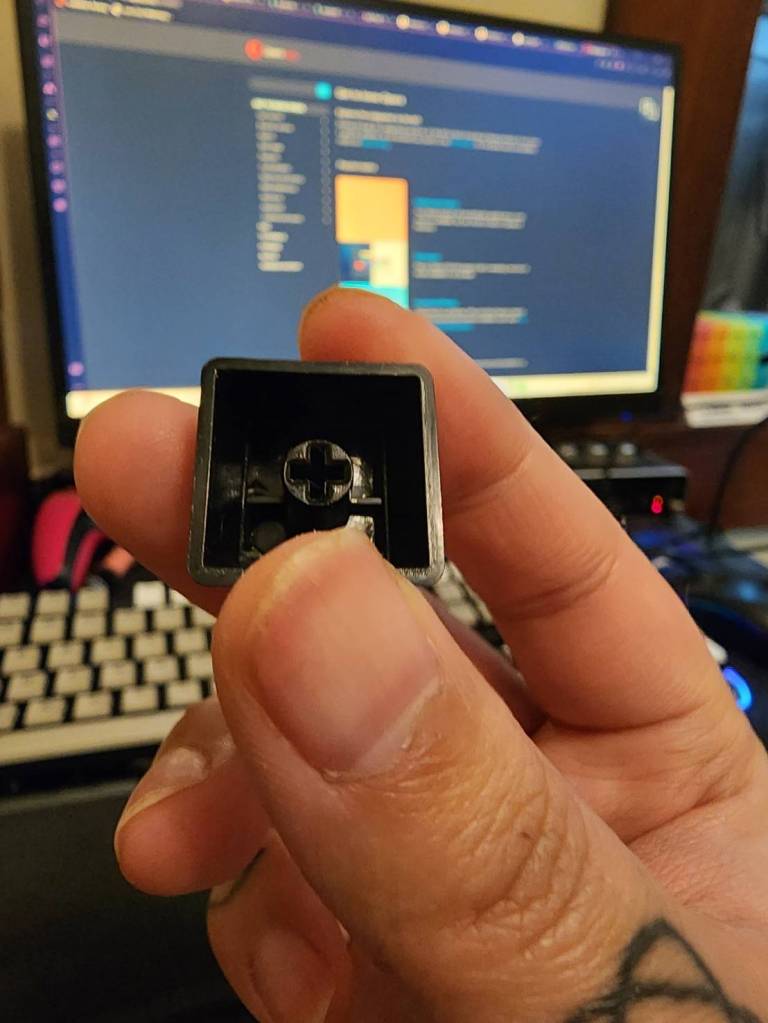 A hand holding the escape keycap from a Hexgears GK735 keyboard, turned upside down to show the underside of the keycap.